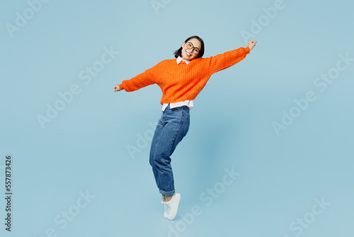 Leinwand Poster Full body side view fun young woman of Asian ethnicity wear orange sweater glasses stand on toes leaning back with outstretched hands dance isolated on plain pastel light blue cyan background studio