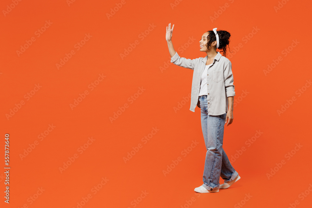 Fototapeta premium Full body sideways profile fun young woman of African American ethnicity she wears grey shirt headband walk go waving hand isolated on plain orange background studio portrait People lifestyle concept