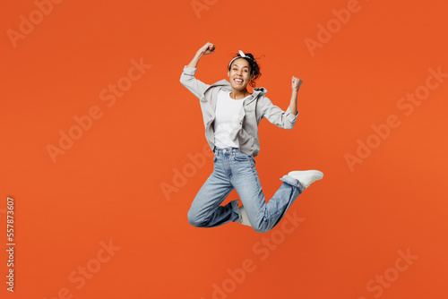 Full body young woman of African American ethnicity she wears grey shirt headband jump high doing winner gesture celebrate clenching fists say yes isolated on plain orange background studio portrait.