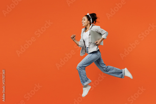 Murais de parede Full body fun side view young IT woman of African American ethnicity she wears grey shirt headband jump high holdclosed laptop pc computer run fast hurrying isolated on plain orange background studio