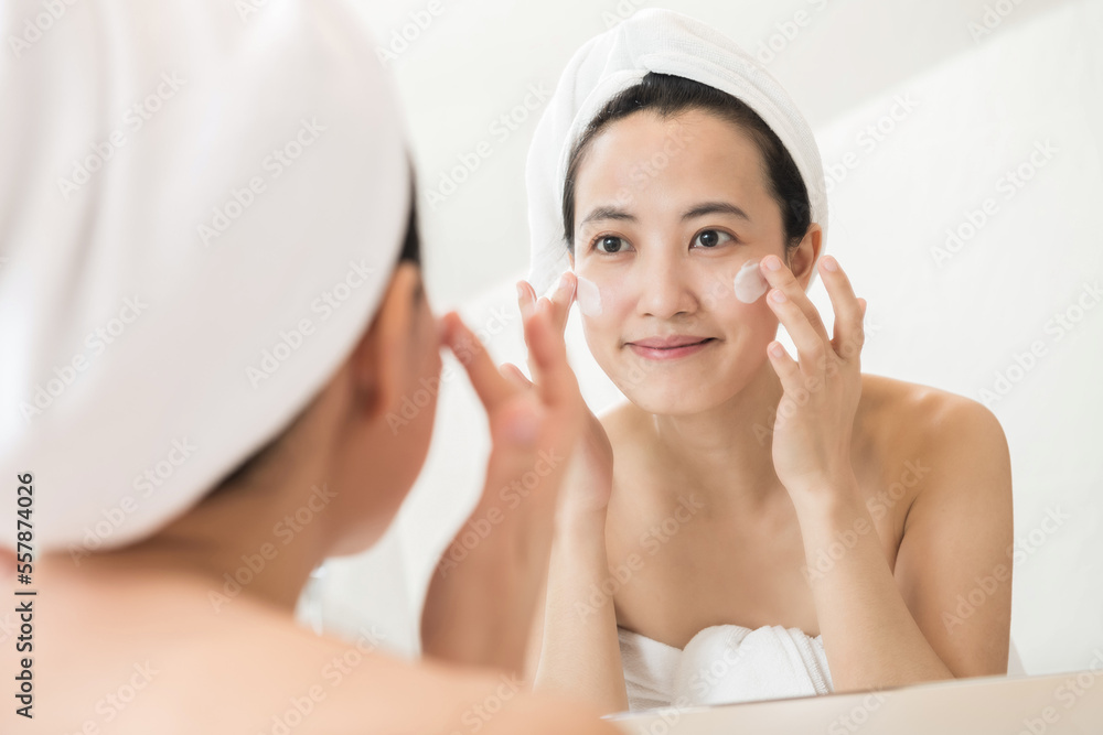 Happy young Asian woman applying face lotions while wearing a towel and touching her face in bathroom