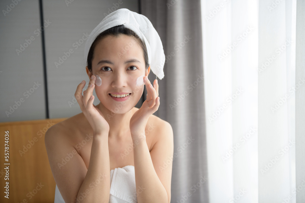 Happy young Asian woman applying face lotions while wearing a towel and touching her face. Daily makeup and skincare Happy young Asian woman applying face lotions while wearing a towel and touching her face. Daily makeup and skincare