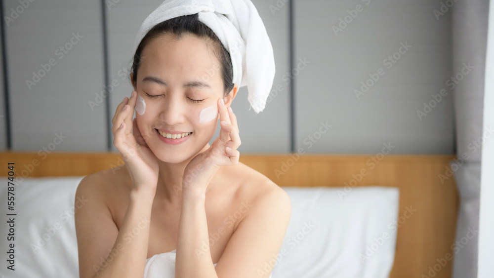Happy young Asian woman applying face lotions while wearing a towel and touching her face. Daily makeup and skincare Happy young Asian woman applying face lotions while wearing a towel and touching her face. Daily makeup and skincare