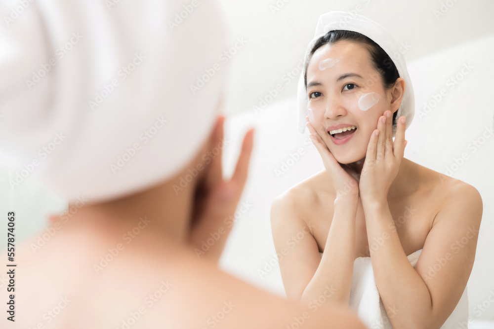 Happy young Asian woman applying face lotions while wearing a towel and touching her face in bathroom Happy young Asian woman applying face lotions while wearing a towel and touching her face in bathroom