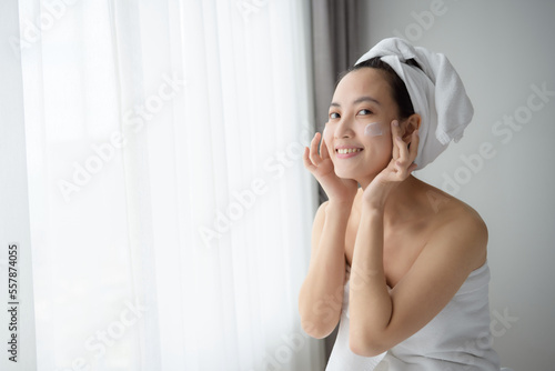 Happy young Asian woman applying face lotions while wearing a towel and touching her face. Daily makeup and skincare