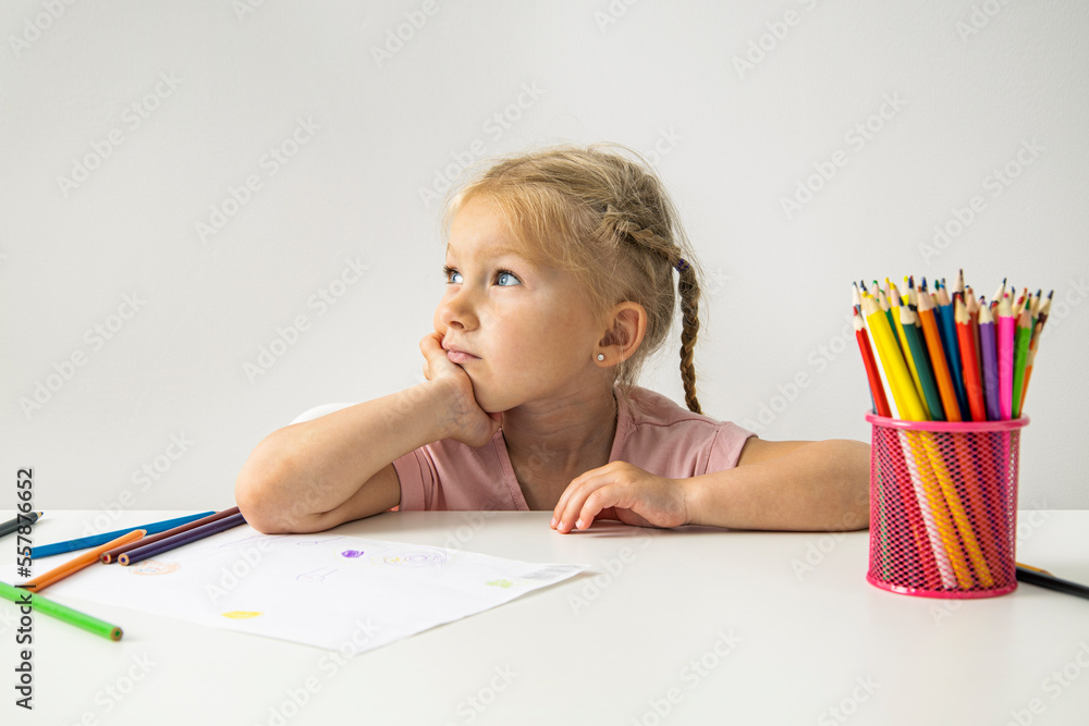 A thoughtful child blonde girl draws with colored pencils sitting at a white table A thoughtful child blonde girl draws with colored pencils sitting at a white table