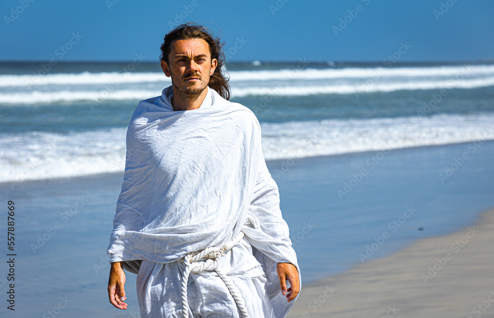 Bearded, handsome man in white robe (resembles Christ) walking next to sea