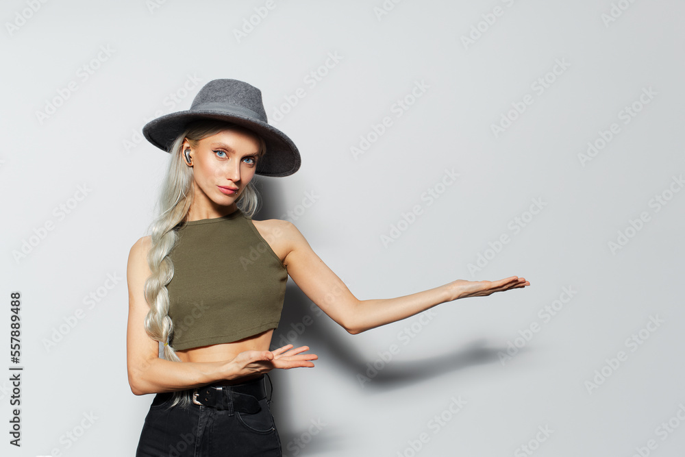 Studio portrait of happy pretty blonde girl with wireless earphones into ears, showing with hands on copy space, wearing grey hat, on white background. Studio portrait of happy pretty blonde girl with wireless earphones into ears, showing with hands on copy space, wearing grey hat, on white background.