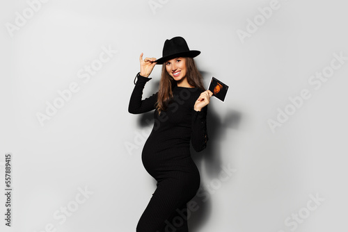 Portrait of joyful pregnant woman, wearing black dress and hat, holding ultrasound scan of her baby.