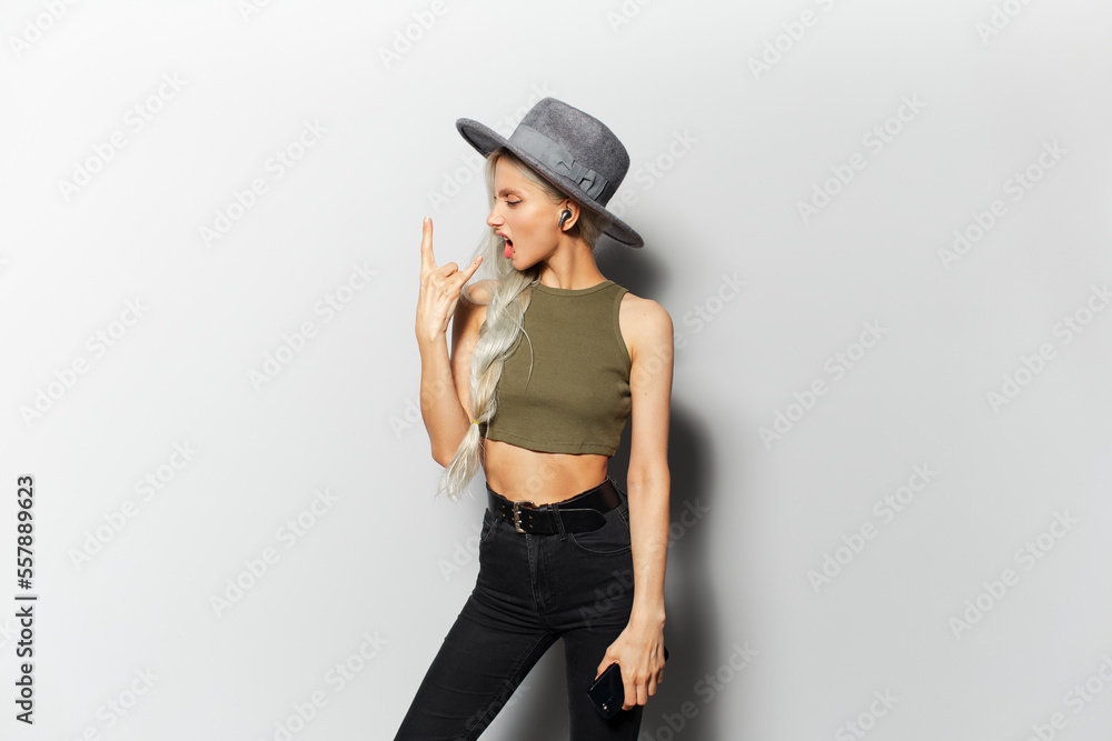 Studio portrait of happy pretty blonde girl with wireless earphones into ears, dancing while listening to music, making rock n roll gesture, wearing grey hat, on white background. Studio portrait of happy pretty blonde girl with wireless earphones into ears, dancing while listening to music, making rock n roll gesture, wearing grey hat, on white background.