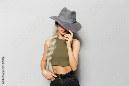 Studio portrait of serious pretty blonde girl with wireless earphones into ears, closes face with grey hat, on white background. © Lalandrew