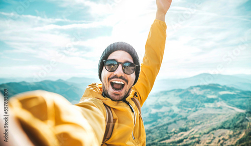 Happy man with backpack taking selfie picture on the top of a mountain - Cheerful hiker climbing cliff - Travel blogger influencer recording adventure live stream on social media platform