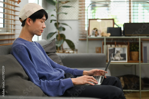 Side view of hipster man surfing internet on laptop while resting on couch in cozy living room