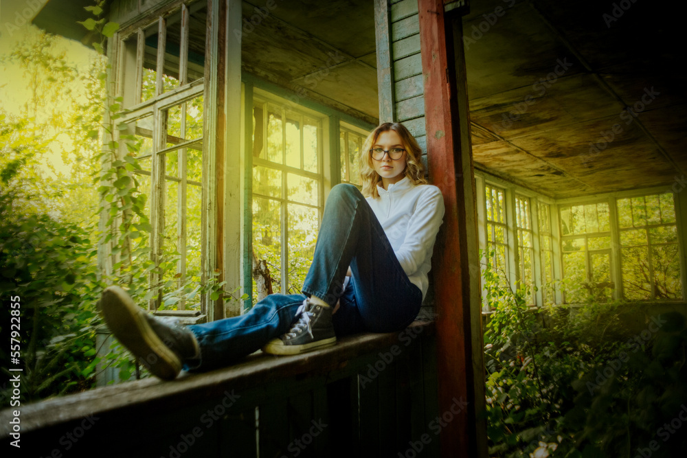 A beautiful blonde woman dressed in a white turtleneck and blue jeans is sitting on a wooden windowsill on the wooden veranda of an old wooden abandoned house on a sunny summer day. Youth vibe. A beautiful blonde woman dressed in a white turtleneck and blue jeans is sitting on a wooden windowsill on the wooden veranda of an old wooden abandoned house on a sunny summer day. Youth vibe.