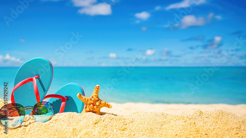 Beautiful colorful background for summer beach holiday. Sunglasses, starfish, turquoise flip-flops on sandy tropical beach against blue sky with clouds on bright sunny day.