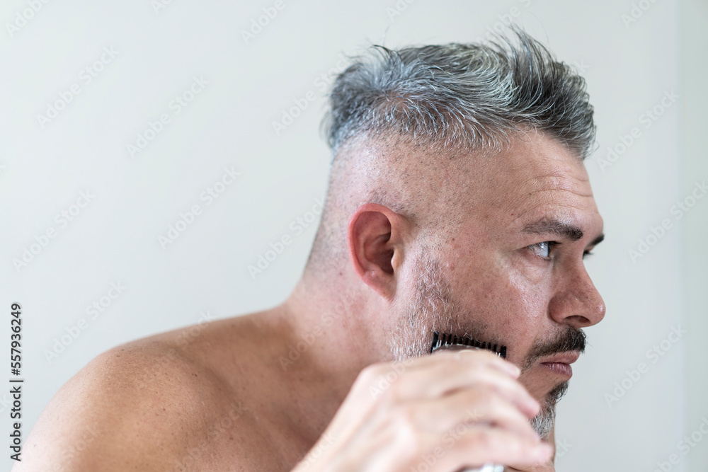 Obraz premium Handsome grizzled bearded man trimming his beard with a trimmer - Middle age man looking himself to mirror and cutting the beard at bathroom - Beauty, hygiene, shaving, grooming concept