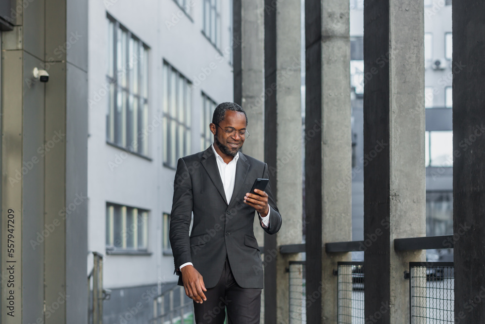 Happy successful African American businessman boss walking outside office building, manager holding smartphone reading message and smiling, using communication app. Happy successful African American businessman boss walking outside office building, manager holding smartphone reading message and smiling, using communication app.