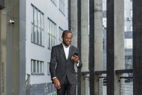 Happy successful African American businessman boss walking outside office building, manager holding smartphone reading message and smiling, using communication app.