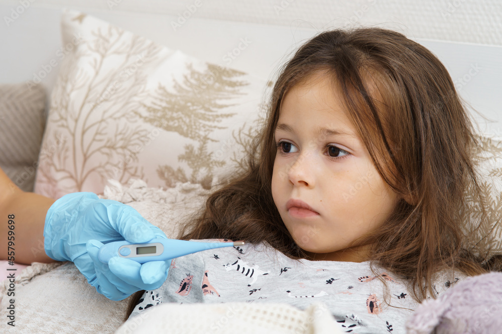 Doctor hand in medical glove measure temperature of sick little girl with electronic thermometer. Portrait of unhealthy child. Kid in sleepwear lie under blanket with fever heat and have bed rest. Doctor hand in medical glove measure temperature of sick little girl with electronic thermometer. Portrait of unhealthy child. Kid in sleepwear lie under blanket with fever heat and have bed rest.