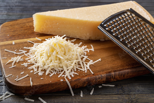 Finely grated parmesan over wooden cutting board. Whole and grated grana padano cheese and stainless steel grater closeup. Italian hard cheese. Delicious dairy product.