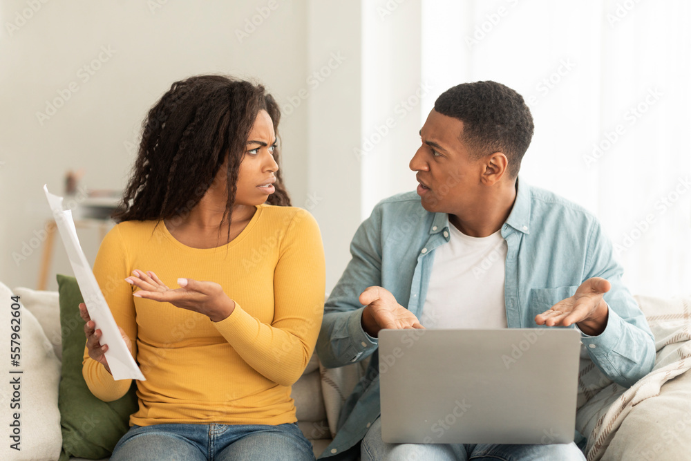 Angry sad millennial african american couple quarrel over debts and bills with papers and laptop in living room Angry sad millennial african american couple quarrel over debts and bills with papers and laptop in living room