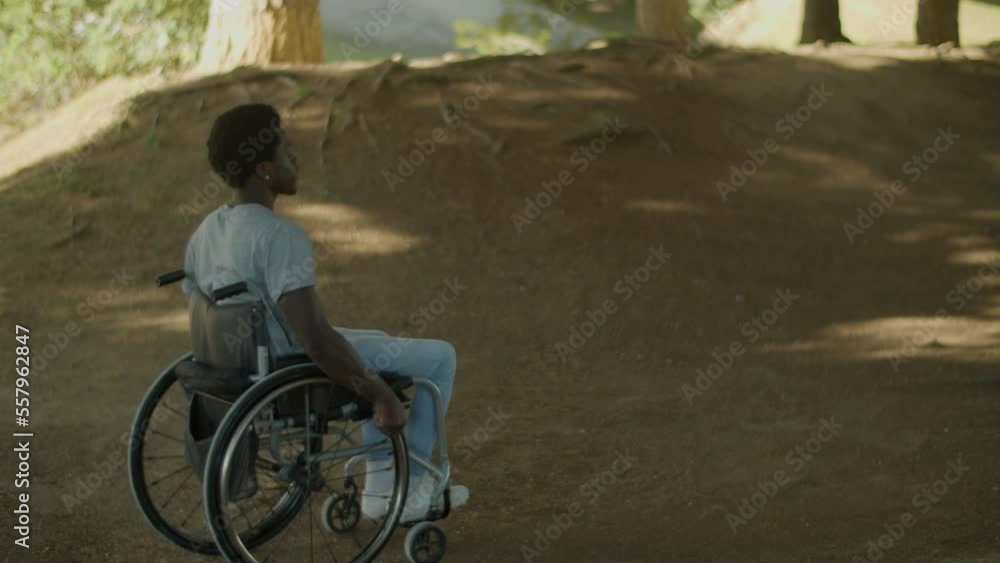 Young Black man riding his wheelchair outside on summer day. Tracking shot of African American guy with disability pushing wheelchair along dirt road. Full shot. Slow motion. Disability concept. Young Black man riding his wheelchair outside on summer day. Tracking shot of African American guy with disability pushing wheelchair along dirt road. Full shot. Slow motion. Disability concept.