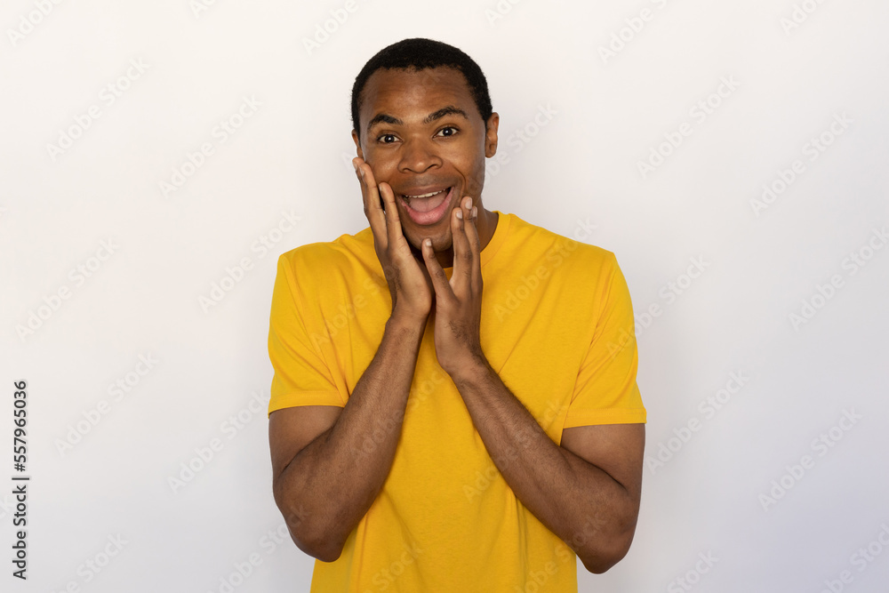 Cheerful young man holding his face with hands from surprise. Male African American model with brown eyes and short black haircut in yellow T-shirt seeing pleasant surprise. Positive emotion concept Cheerful young man holding his face with hands from surprise. Male African American model with brown eyes and short black haircut in yellow T-shirt seeing pleasant surprise. Positive emotion concept