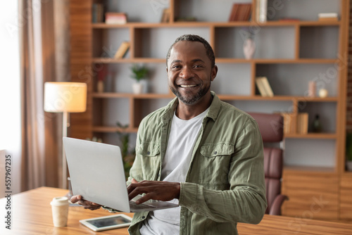 Smiling attractive mature black male manager with laptop work, make presentation and look at camera