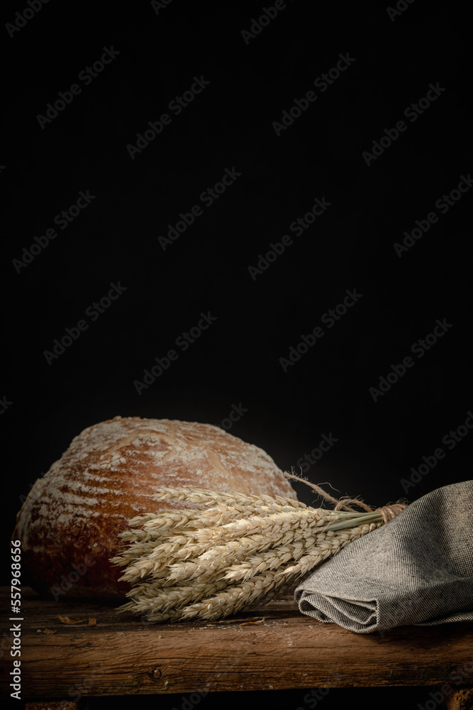 Still life with freshly baked bread and a sheaf of wheat ears on the rustic wooden table with copy space on black background.