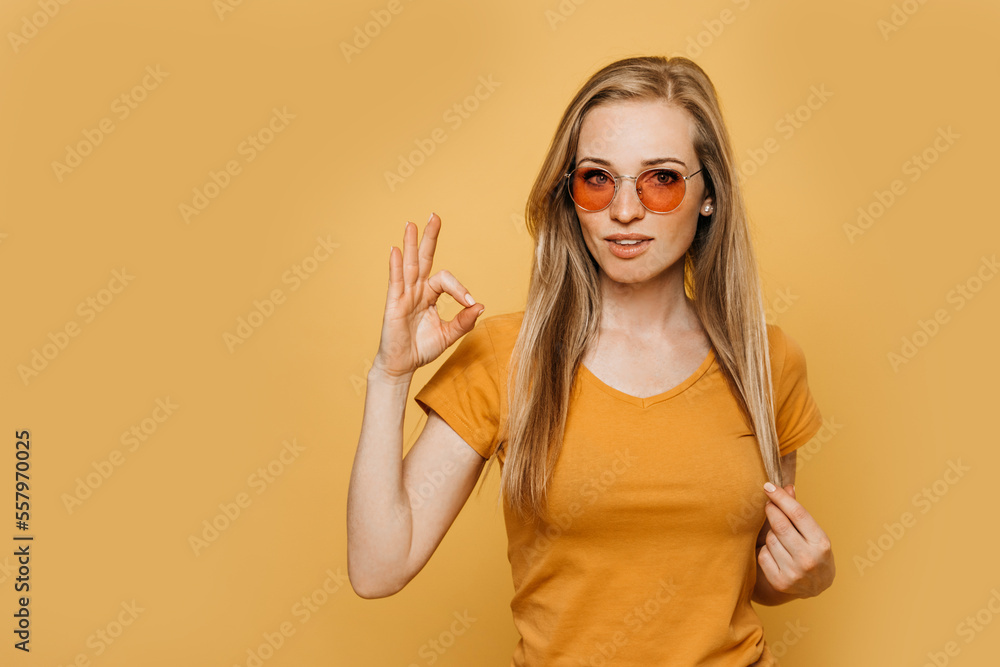 Charming redhead girl in orange sunglasses, dressed in orange t-shirt, looks confident, makes ok gesture, approves your choice, over yellow background. Trust me, choose it.