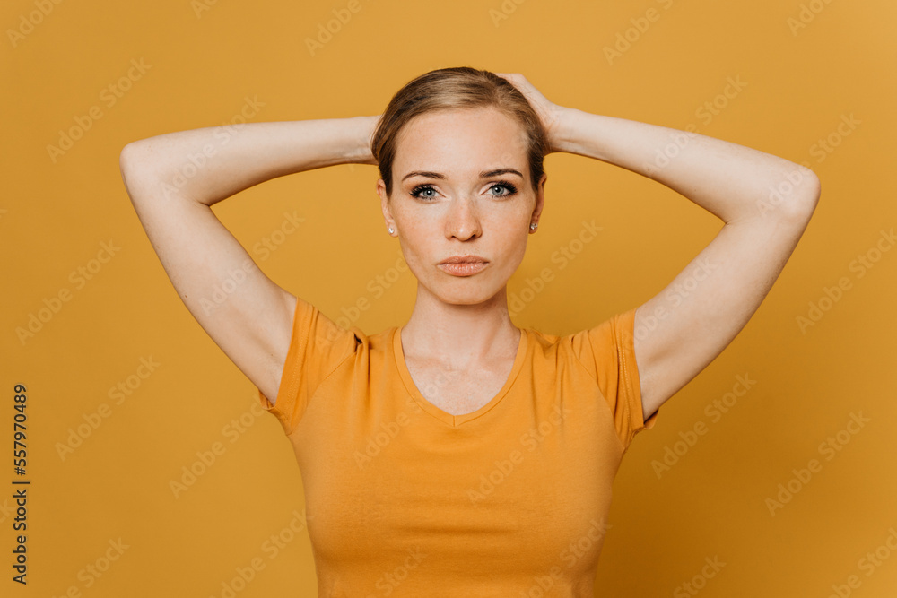 Beautiful young red haired woman in orange t-shirt, collects her hair on the head by both hands, looks confident, over yellow background. Purposeful people concept. Beautiful young red haired woman in orange t-shirt, collects her hair on the head by both hands, looks confident, over yellow background. Purposeful people concept.