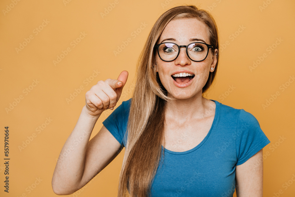 Close up portrait of cheerful excited young woman in glasses with open mouth, dressed in blue t-shirt, pointing finger at camera isolated on the yellow backdrop with copy space. Active people concept.