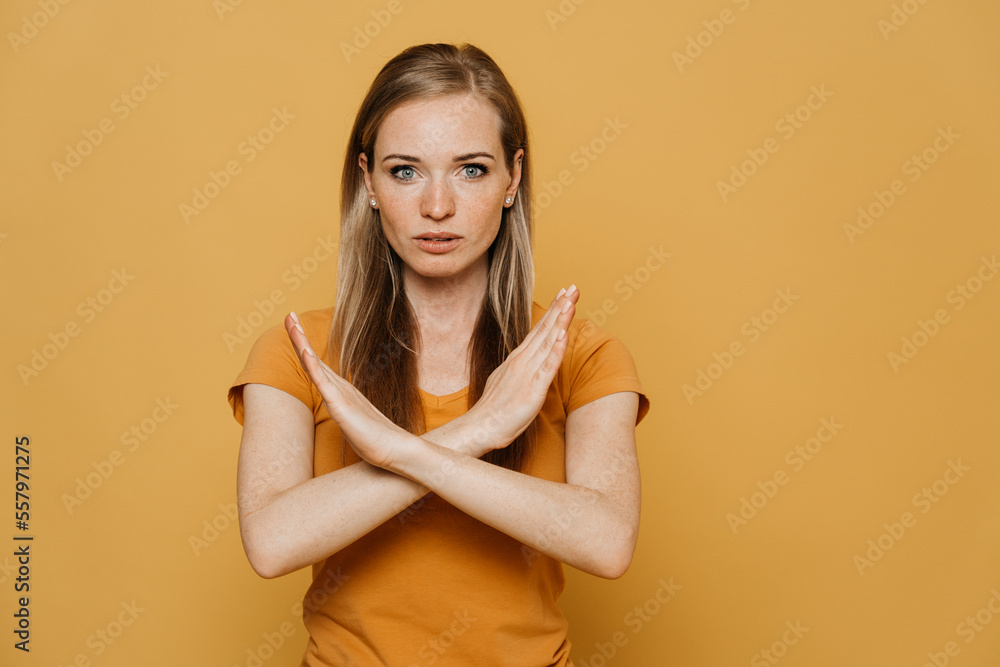 Obraz premium Confident redhead pretty woman in orange t-shirt, shows gesture sign stop, crossing her arms, wants to stop the coronavirus pandemic. Stay home and health care concept. Stands over yellow background.