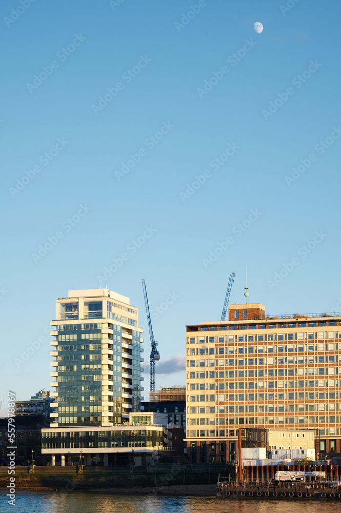 Tall building in sunshine with crane in front of river thames in London and moon overhead