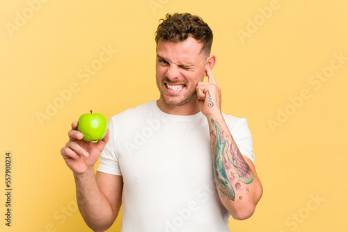 Young caucasian man eating an apple isolated covering ears with hands.