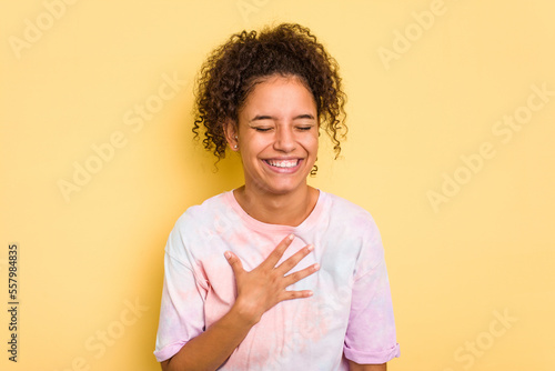 Young Brazilian curly hair cute woman isolated on yellow background laughs out loudly keeping hand on chest.