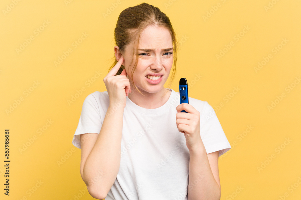 Young cute woman holding a vaper isolated on yellow background covering ears with hands. Young cute woman holding a vaper isolated on yellow background covering ears with hands.