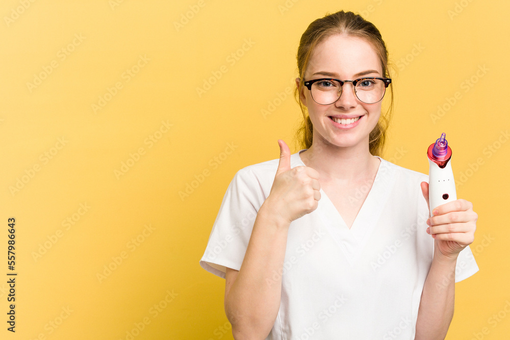 Young beautician woman holding a facial cleaner isolated on yellow background smiling and raising thumb up Young beautician woman holding a facial cleaner isolated on yellow background smiling and raising thumb up