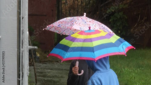 Two anonymous unrecognizable school age girls, children standing in the rain holding umbrellas together outdoors, simple scene. Sad rainy weather day, raining outside, closeup, slow motion, lifestyle