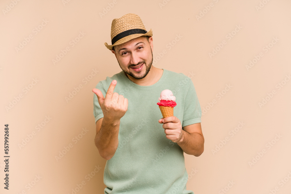 Young latin man holding an ice cream isolated pointing with finger at you as if inviting come closer. Young latin man holding an ice cream isolated pointing with finger at you as if inviting come closer.