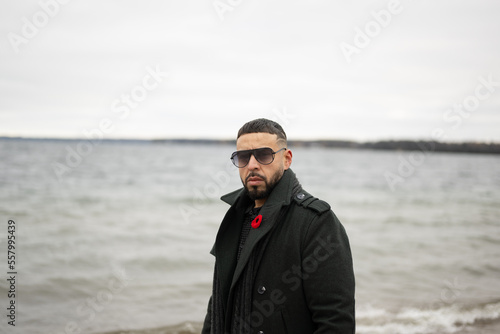 Bi racial model standing on board walk with water behind him winter time with sunglasses and green jacket on 