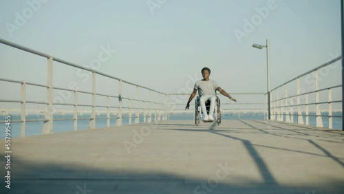 Front view shot of Black man riding wheelchair at seafront. Young man with paraplegia enjoying sunny day at seashore, pushing wheels, approaching camera. Low angle shot. Motivation, disability concept