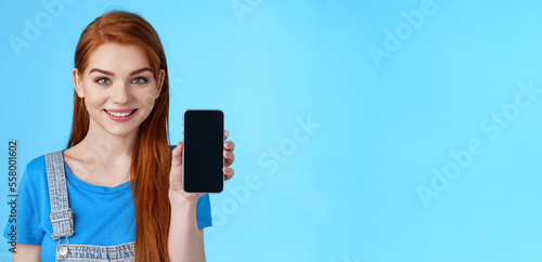 Close-up attractive joyful redhead young 20s female shop assitant suggest customer buy smartphone, show phone display, promote application, internet app, smiling camera, stand blue background photo