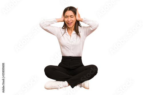 Young colombian woman sitting on the floor isolated covering ears with hands trying not to hear too loud sound. © Asier