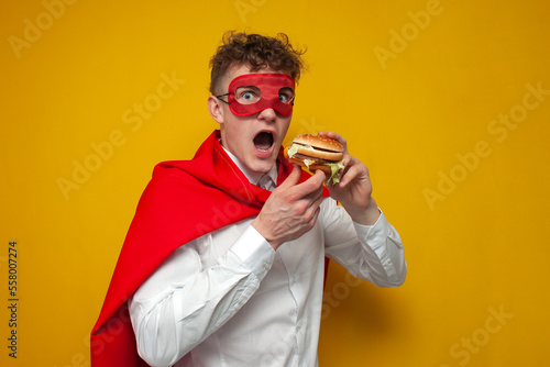 young guy in a superhero costume eats a delicious burger on a yellow background, super man eats fast food