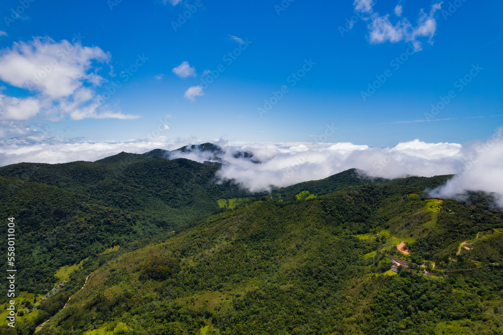 Naklejka premium Aerial image of mountains with low clouds covering part of the landscape. Heavy clouds, green vegetation and very blue sky. Mist and white clouds.