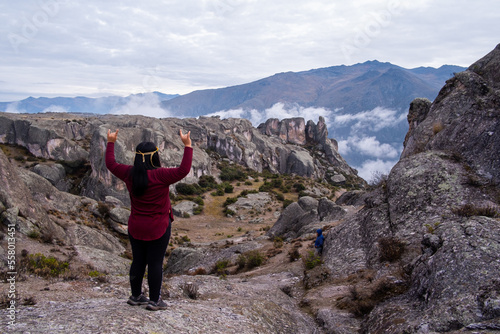 Latin woman with her back turned and her arms raised, making the sign of the metal, standing on the edge of a rock, looking towards the valley.