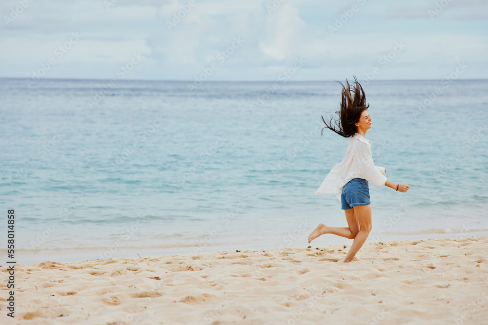 Naklejka premium Sports woman runs along the beach in summer clothes on the sand in a yellow T-shirt and denim shorts white shirt flying hair ocean view, beach vacation