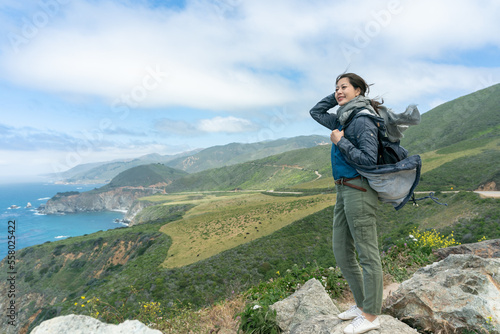 full length of cheerful asian taiwanese woman hiker standing on rock near cliff and enjoying scenic view of mountain and sea in the wind at big sur california usa