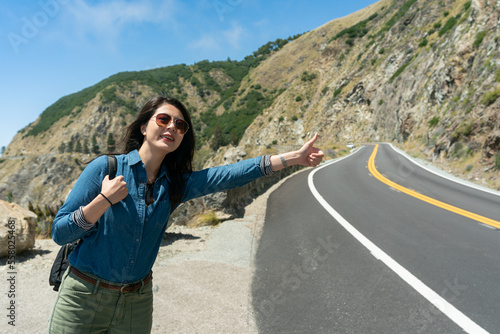 smiling asian Chinese female traveler on hiking road trip hitchhiking on road at mountainside in California usa